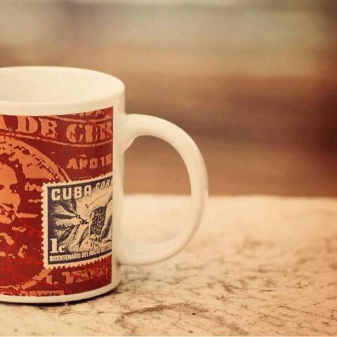 A white coffee mug decorated with a vintage-style Cuban postage stamp graphic sits on a textured surface.